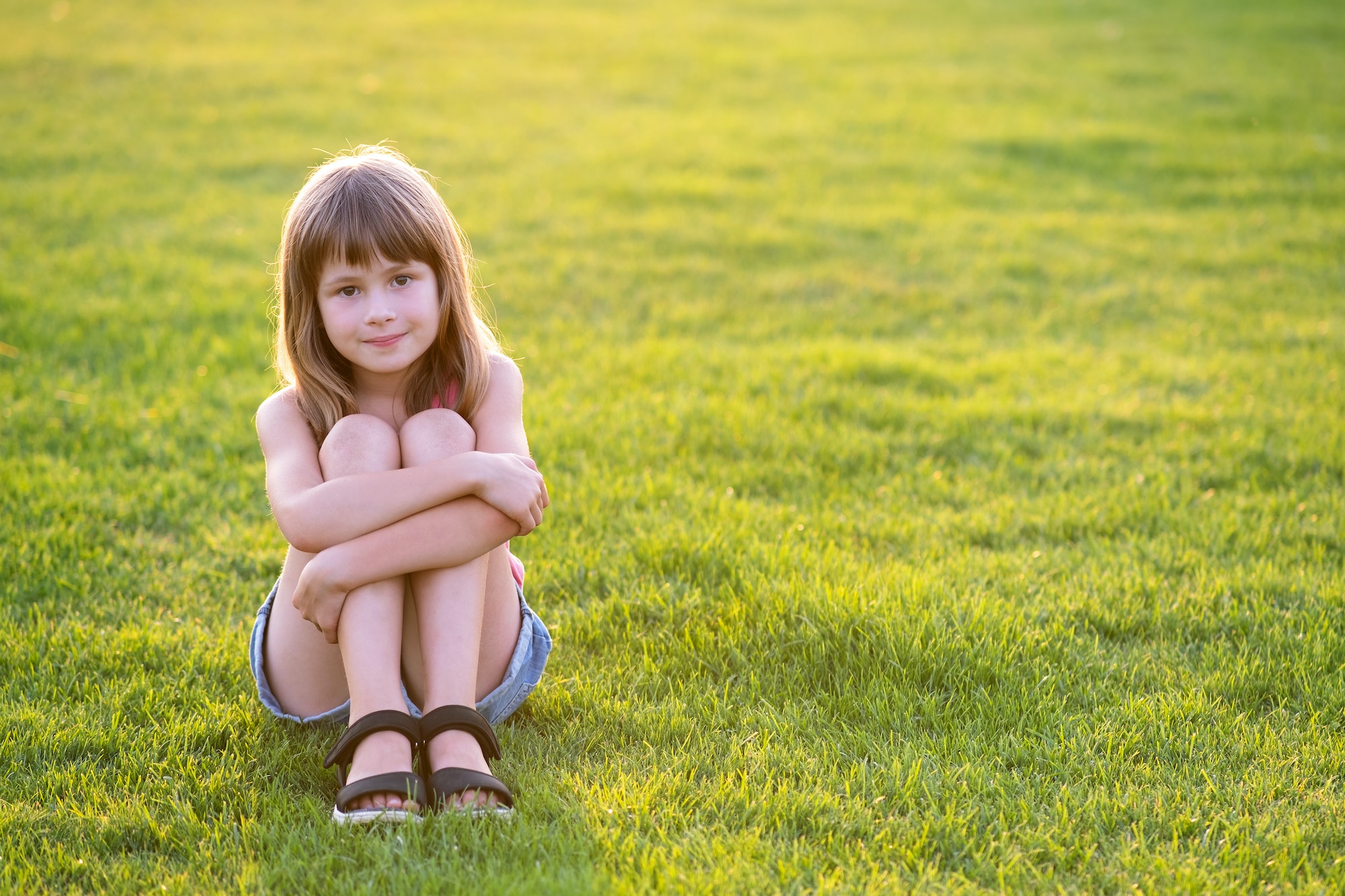 Young happy child girl resting while sitting on green grass lawn on warm summer day